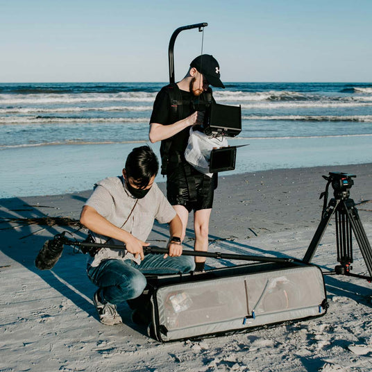 Two people setting up filming equipment on a beach with ocean waves in the background