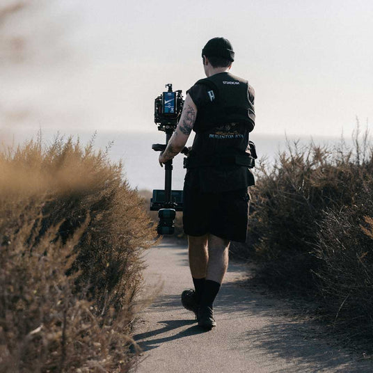 Person with filming equipment walking on a path through dry bushes